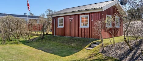 Un edificio de madera roja con dos ventanas y un pequeño porche, rodeado de césped y árboles.