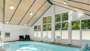 En indendørs swimmingpool med glasvægge, træbjælker i loftet og udsigt til træer.