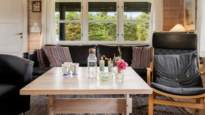 A cozy room with a wooden ceiling, a large window with a view of greenery, a table with a vase of flowers, and a black leather chair.