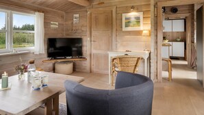 A cozy cabin interior with a wooden ceiling, a TV on a stand, a dining table with chairs, and a kitchen area.