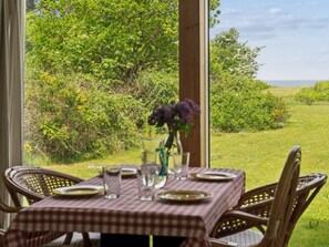 Un espace de restauration en plein air avec une table dressée pour quatre personnes, entourée de chaises en osier, donnant sur un paysage verdoyant avec des arbres et une vue sur la mer.