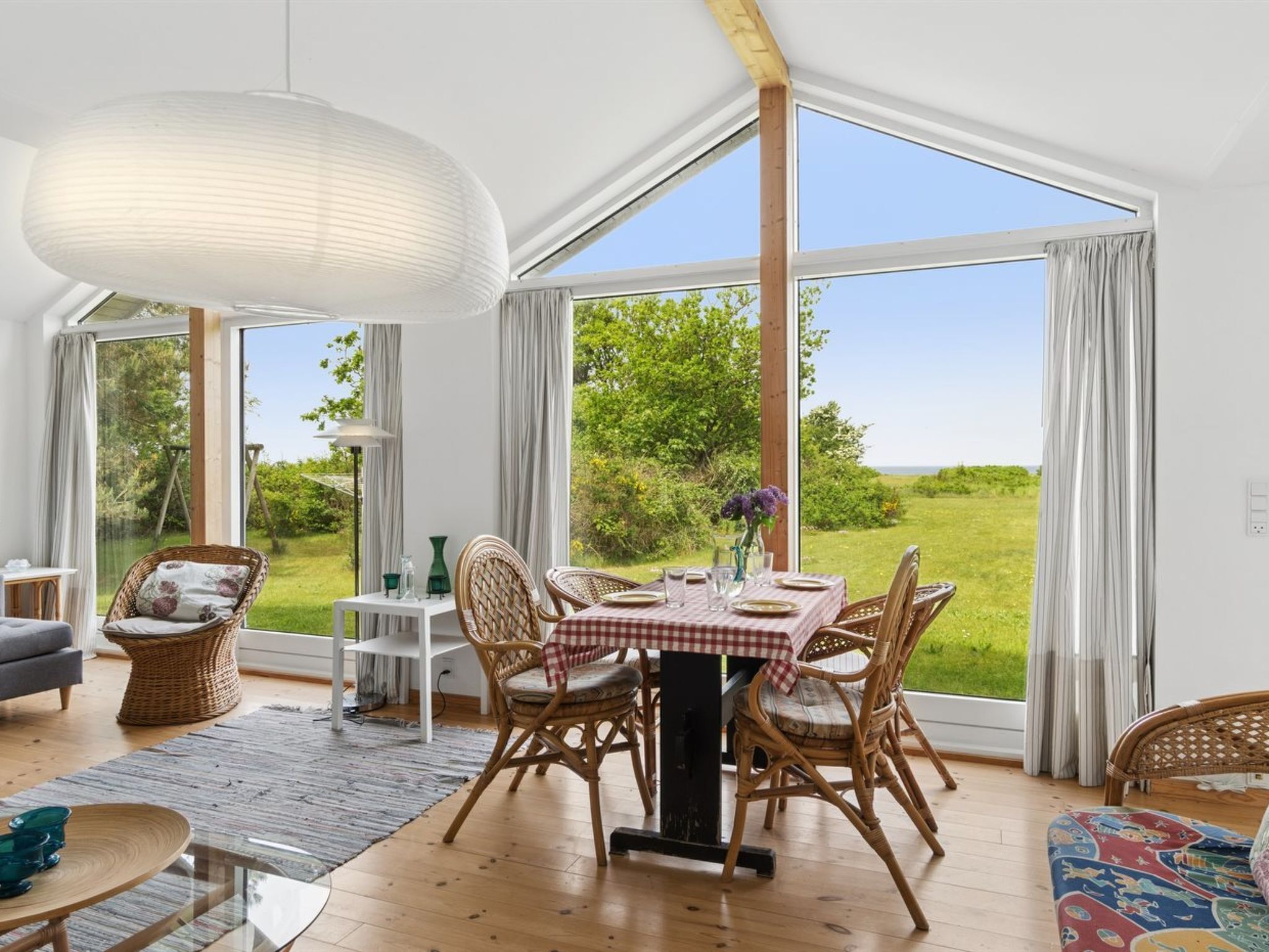 Een eethoek met een tafel voor vier personen, omringd door rieten stoelen, een groot raam met uitzicht op groen en een hanglamp.