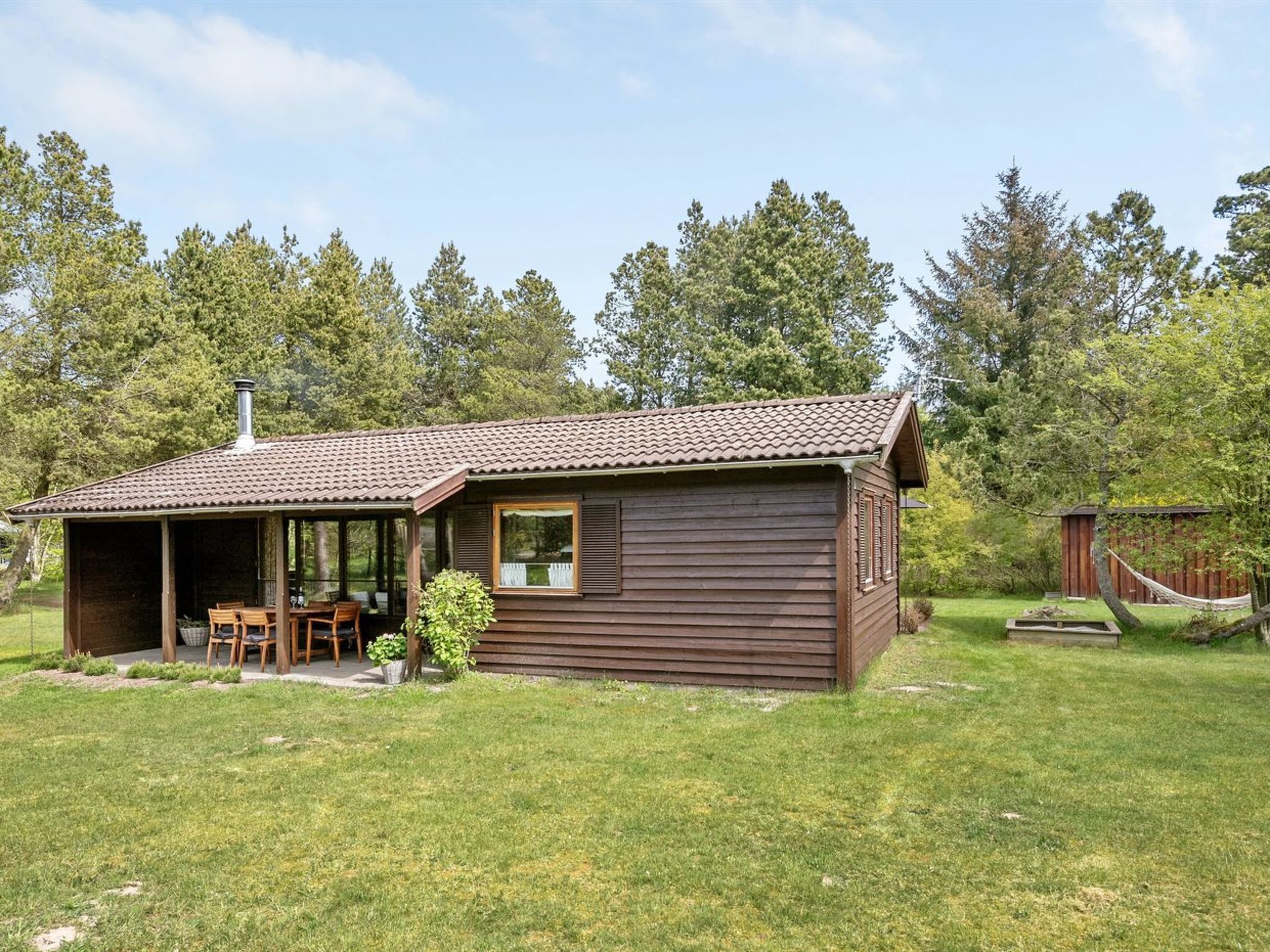 Une cabane en bois avec un toit en tuiles, une véranda et un petit jardin agrémenté d’une table et de chaises.