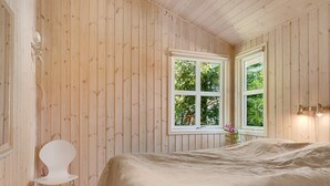 Une chambre avec des murs en bois, un lit recouvert d’une couvre-lit à motifs, une chaise blanche et deux fenêtres donnant sur un paysage verdoyant.