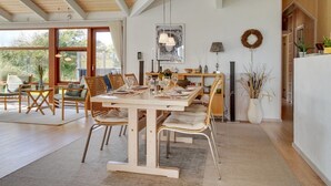 Ein geräumiger Essbereich mit einem Holztisch, gedeckt für sechs Personen, umgeben von Korbstühlen. Ein großes Fenster sorgt für natürliches Licht, und eine rustikale Decke mit sichtbaren Holzbalken ist zu sehen.