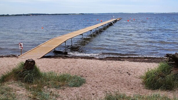 Eine Holzstege ragt in ein Gewässer mit einem sandigen Strand im Vordergrund.