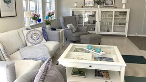 Ein Wohnzimmer mit einem weißen Sofa, gemusterten Sesseln, einem Couchtisch mit Zeitschriften und einer weißen Kommode mit Glastüren.