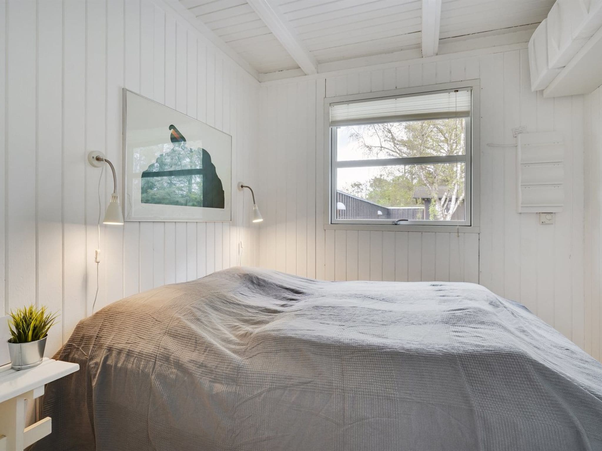 A bedroom with a bed covered in a grey blanket, a bedside table with a plant, a framed picture on the wall, and a window with a view of trees and a building outside.