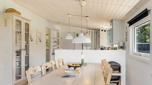 Comedor con una mesa larga, sillas e iluminación colgante. Incluye una zona de cocina con armarios y una ventana.