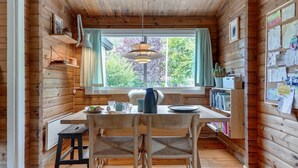 A cozy cabin interior with a wooden dining table, chairs, a hanging light fixture, and a window with a view of trees.