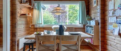 L’intérieur chaleureux d’un chalet avec une table à manger en bois, des chaises, un luminaire suspendu et une fenêtre donnant sur la forêt.