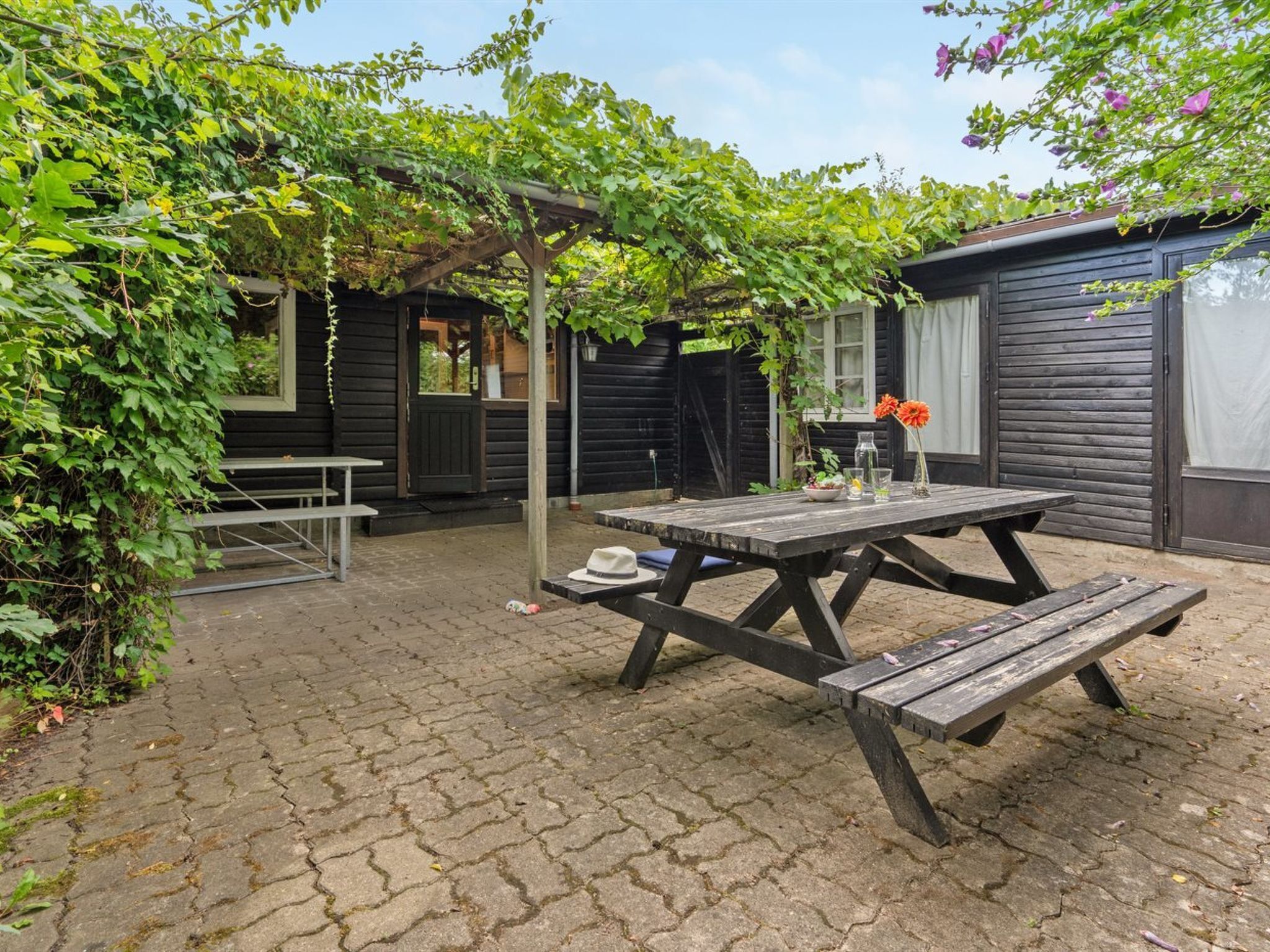 Ein Holztisch mit Bänken in einem Garten mit Backsteinpflaster, umgeben von Grünpflanzen und einem Holzgebäude.