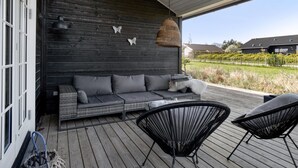 Un espace extérieur abrité, aménagé avec un mur en bois foncé, deux chaises noires et un canapé gris agrémenté de coussins.