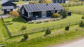 Une maison moderne dotée d’un grand porche, entourée d’une cour clôturée et d’autres maisons similaires, le tout situé dans un cadre rural.