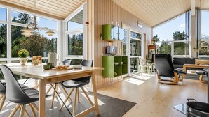 Una habitación amplia con techo de madera, ventanas grandes, zona de comedor con mesa y sillas, un armario verde y un área de descanso con sofás y una mesa de centro.