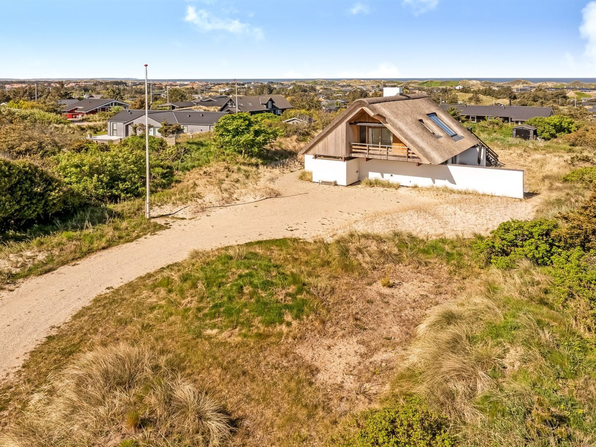 A modern house with a sloped roof, situated on a sandy hill with a view of other houses in the background.