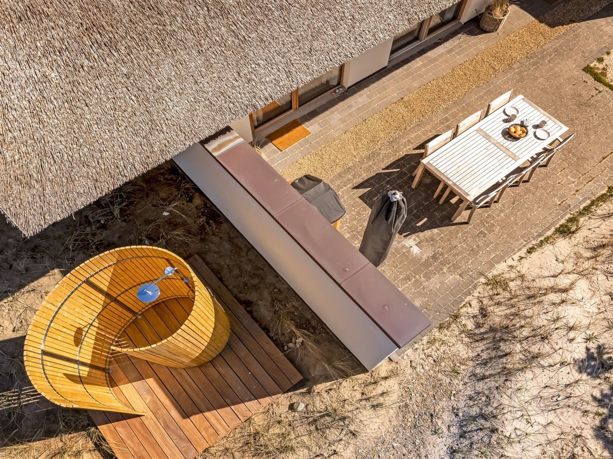Ein überdachter Außenbereich mit Strohdach, einer Holzterrasse, einem Tisch mit Stühlen und einer kleinen Holzkonstruktion.