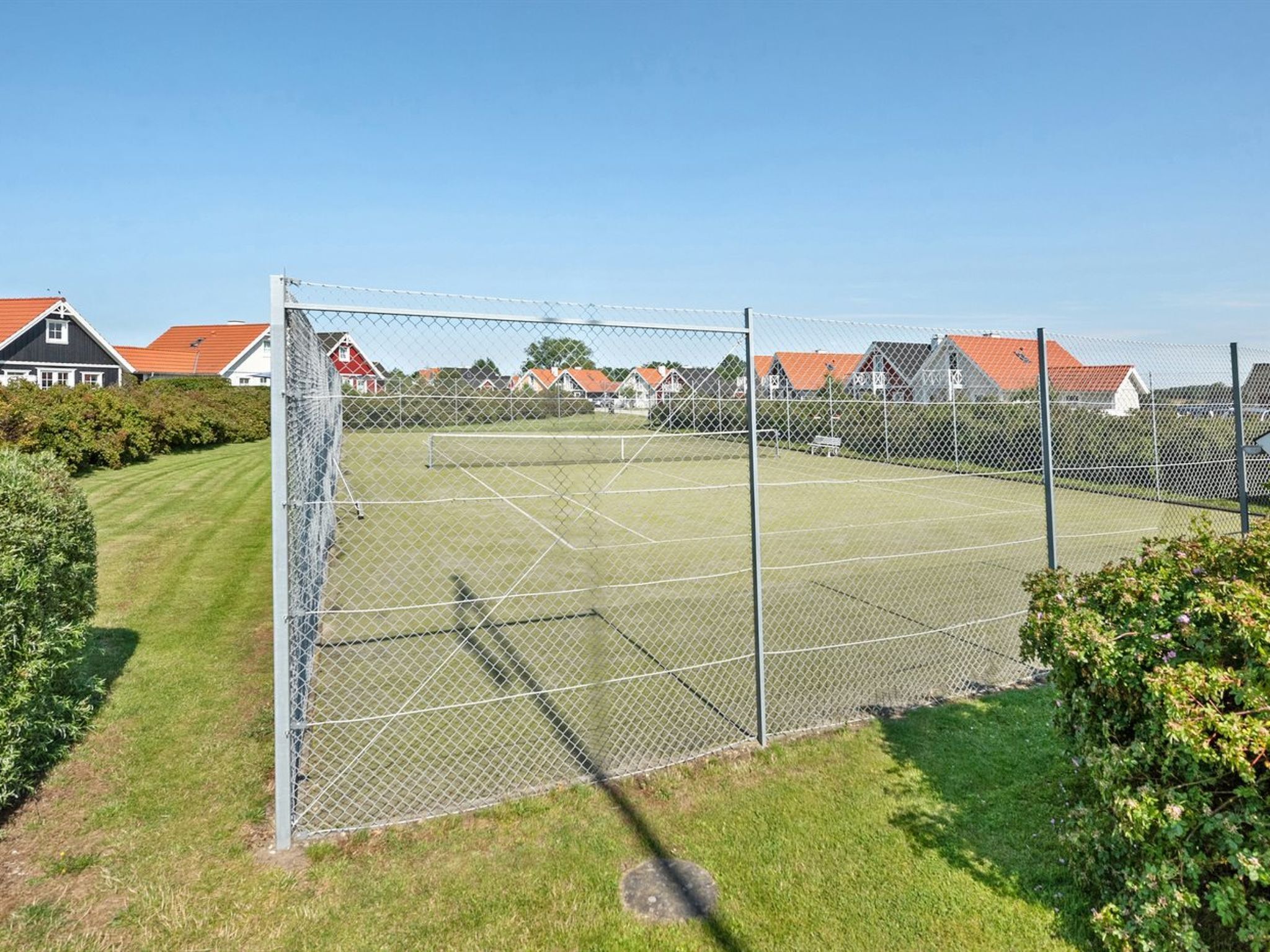 A tennis court enclosed by a chain-link fence, surrounded by well-maintained grass and hedges, with houses in the background.