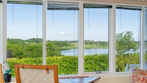 Una habitación con una mesa y sillas de madera, ventanas amplias con vistas a un lago y vegetación.