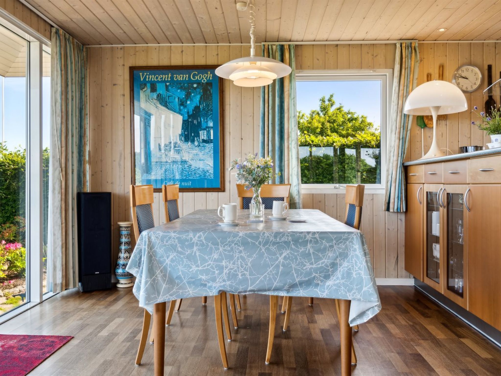 A dining area with a table set for four, wooden paneling, a large poster of Vincent van Gogh, and a view of greenery outside.