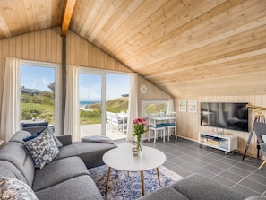 A spacious living area with a large sofa, a round coffee table, a dining table with chairs, a flat-screen TV, and a bookshelf.