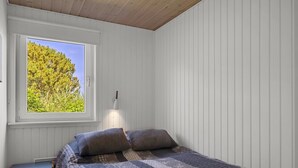 Een slaapkamer met een bed, nachtkastje, raam met uitzicht op bomen en een wandlamp.