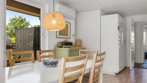 Een moderne keuken met witte kastjes, een eettafel met vier houten stoelen, een geweven hanglamp en uitzicht op de natuur.