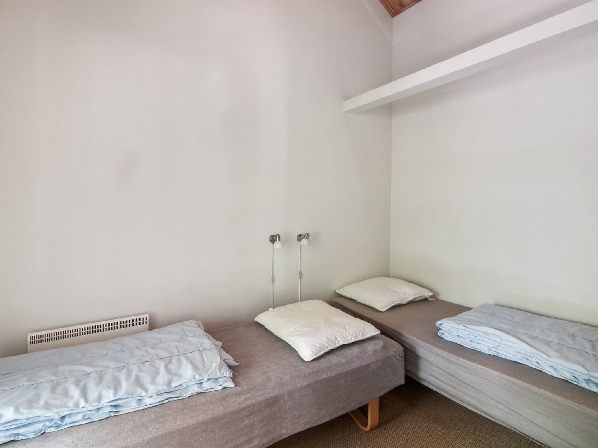 Um alojamento pequeno, com um quarto e duas camas individuais, uma parede branca e uma estante branca.