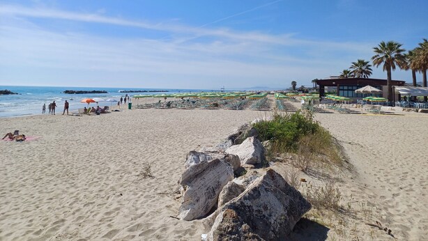 Une plage avec des chaises longues et des parasols, un bâtiment orné d’un palmier, et un premier plan rocheux.