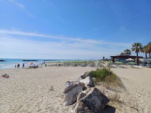 Une plage avec des chaises longues et des parasols, un bâtiment orné d’un palmier, et un premier plan rocheux.