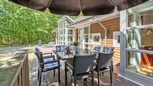 Terraza exterior con mesa de vidrio y sillas de mimbre, rodeada por una cubierta de madera y una casa con ventanales amplios.