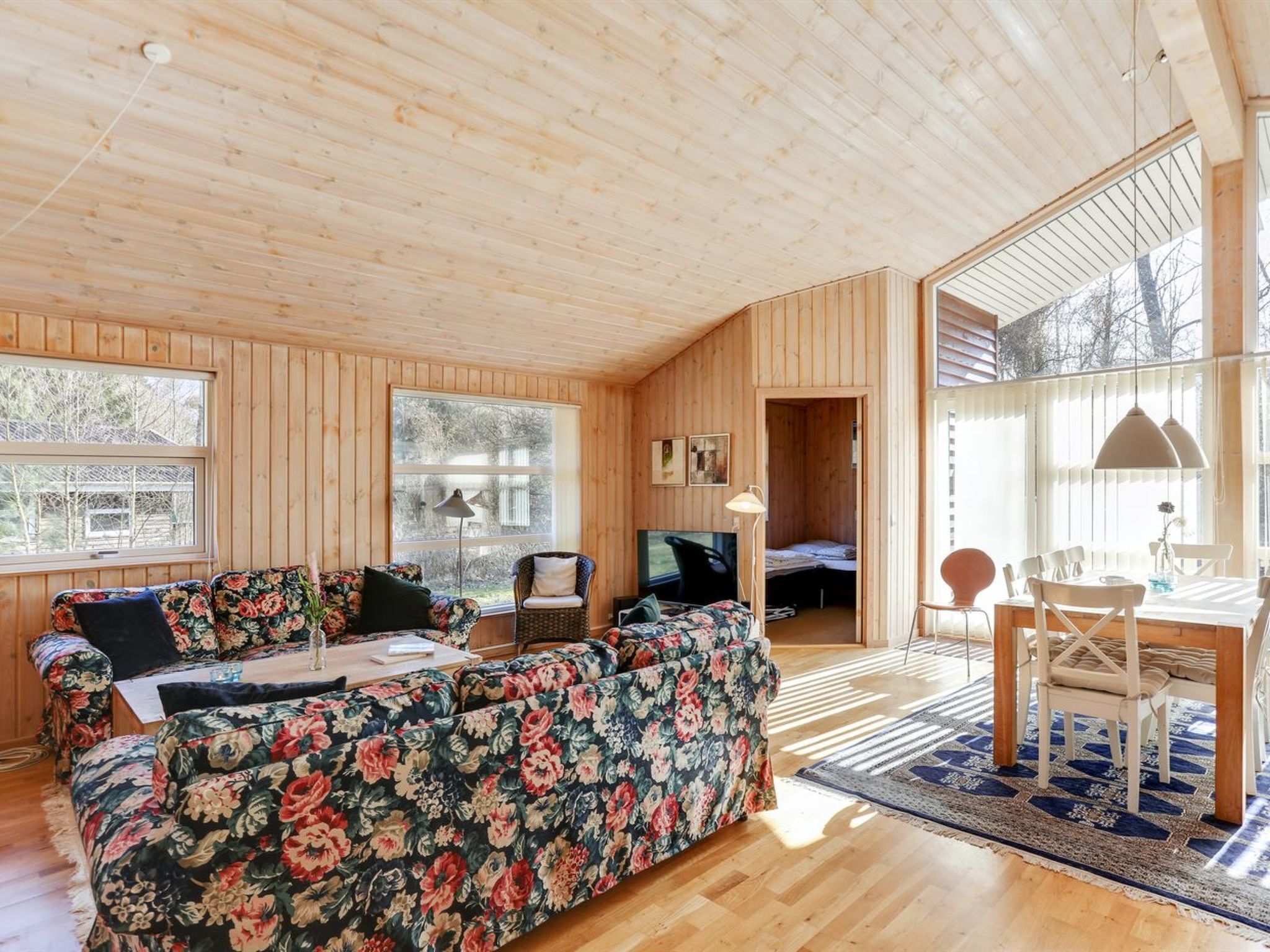 L’intérieur chaleureux d’un chalet avec un plafond en bois, un canapé à motifs floraux, une table à manger avec des chaises et un téléviseur dans un coin.
