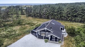 A modern house with a sloped roof, surrounded by dense forest and a gravel driveway.