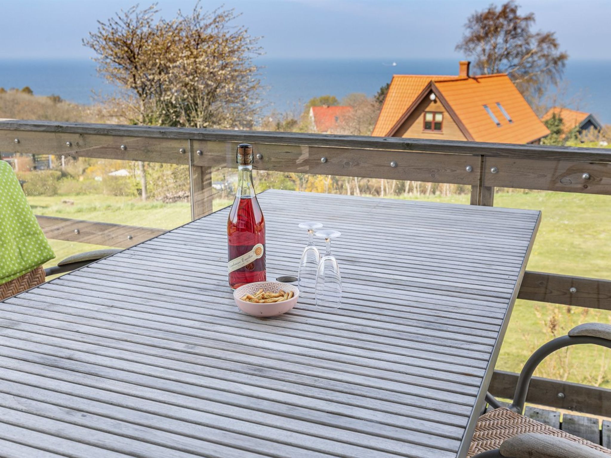 Et bord med en flaske rødvin, en skål med snacks og tre vinglas på en metalterrasse med udsigt til huse og havet i baggrunden.