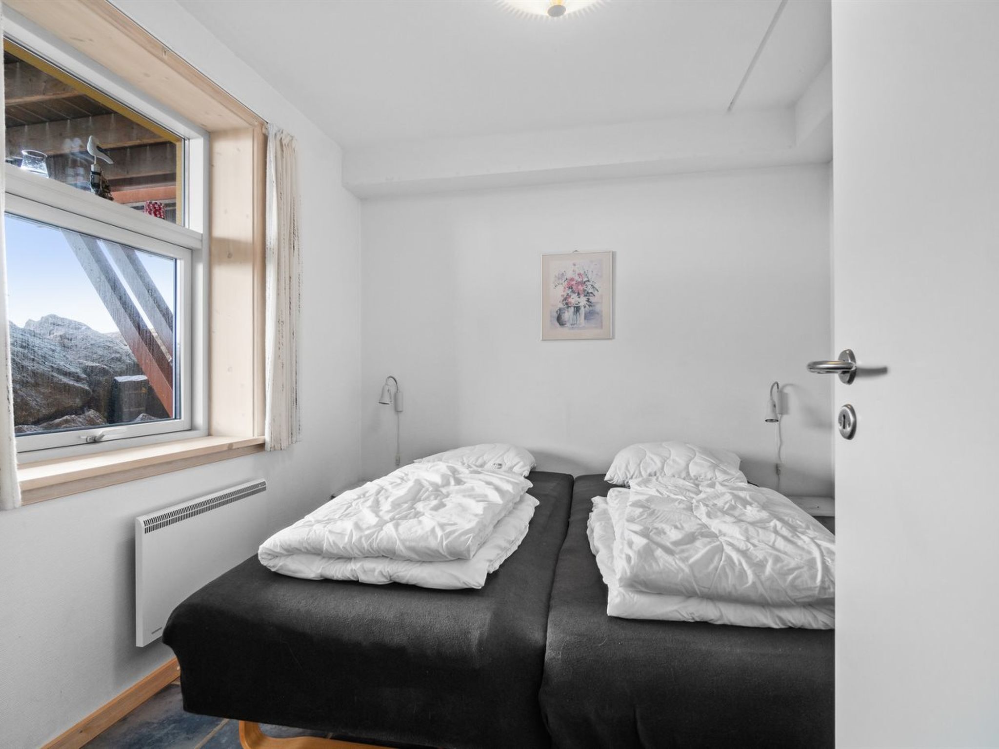 A small, clean room with two beds, a window with a view of mountains, a radiator, and a door with a handle.