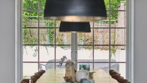 Una mesa de comedor con sillas, una lámpara y vistas a un jardín a través de una ventana con diseño de cuadrícula.