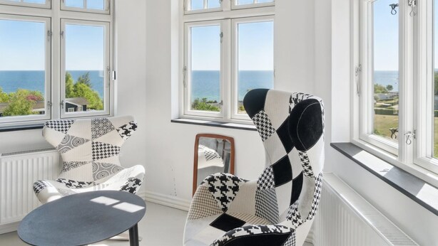 Una habitación con dos ventanas amplias que dan al mar, un sillón estampado, una mesita y un radiador.