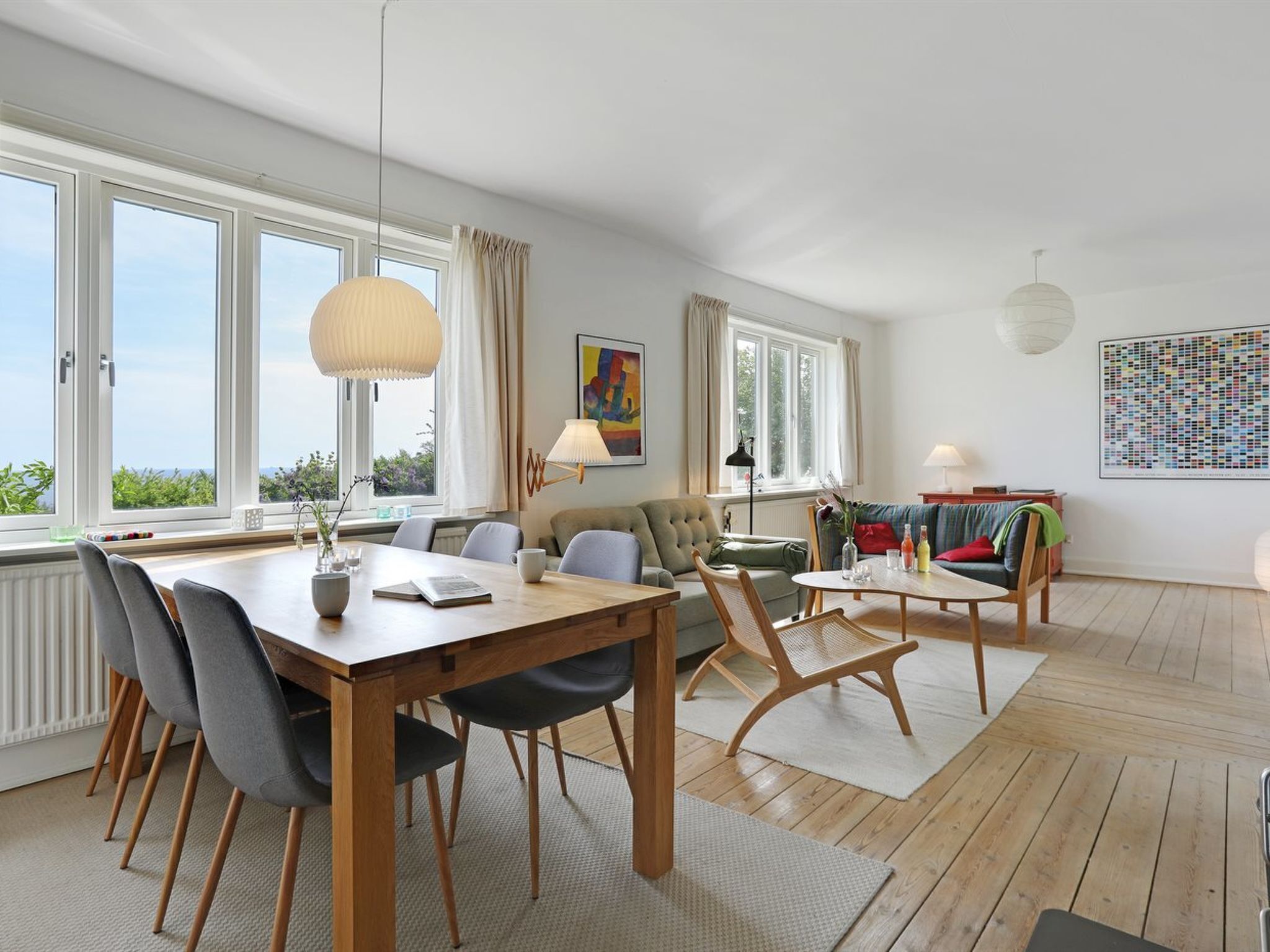 A spacious living room with a wooden dining table, comfortable seating, large windows, and a view of greenery.