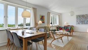 Ett rymligt vardagsrum med ett matbord i trä, bekväma sittplatser, stora fönster och utsikt över grönområden.