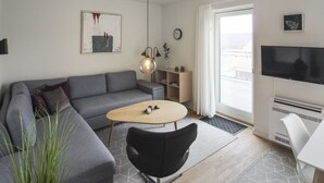 A modern living room with a grey sofa, a round coffee table, a black chair, a TV, and a large window with curtains.
