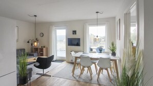 A modern living room with a sofa, dining table, and chairs. There is a TV mounted on the wall, a plant, and a window with curtains.