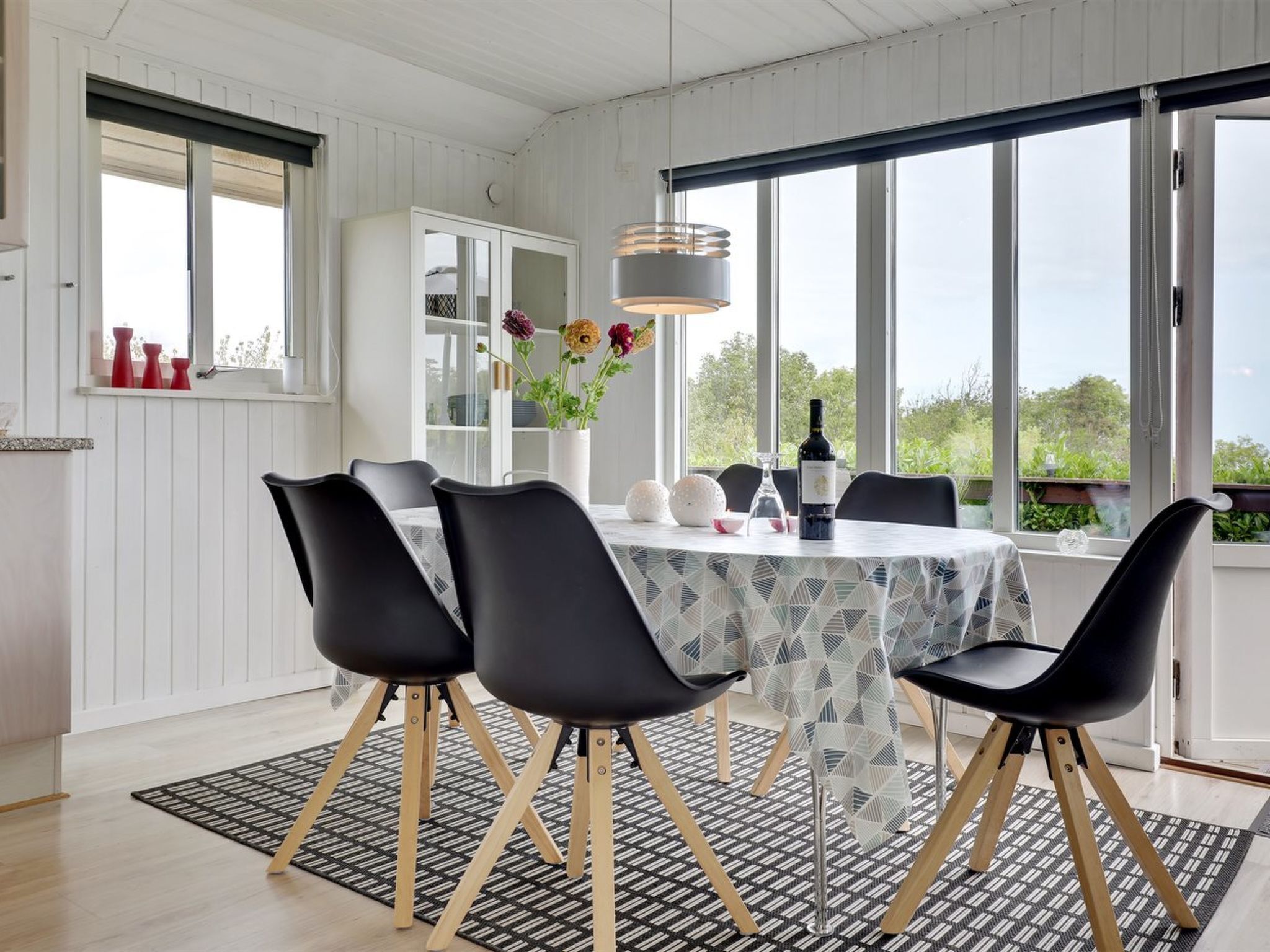Ein Essbereich mit einem runden Tisch, schwarzen Stühlen, einer gemusterten Tischdecke, einer Vase mit Blumen, einer Weinflasche und Blick ins Grüne durch große Fenster.