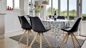 En matplats med ett runt bord, svarta stolar, en mönstrad duk, en vas med blommor, en vinflaska och utsikt över grönskan genom stora fönster.