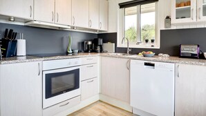 Ett modernt kök med vita skåp, en inbyggd ugn, en diskmaskin och ett fönster med utsikt över grönområden.