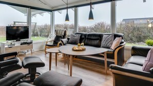 Ein modernes Wohnzimmer mit einer schwarzen Ledersofa, einem runden Couchtisch und einem Fernseher auf einer Holzkommode. Große Fenster bieten einen Blick ins Freie.