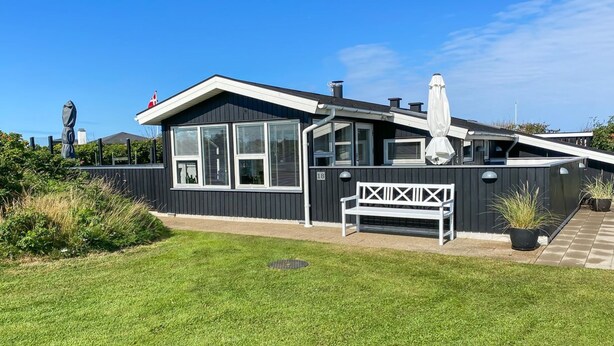 Une maison moderne avec une façade noire, des bordures blanches et de grandes fenêtres. Un banc blanc se trouve devant la maison.