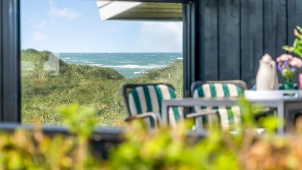 Een kamer met een groot raam dat uitkijkt op een grasveld en de zee. Er staan twee stoelen en een tafel met een bloevas.