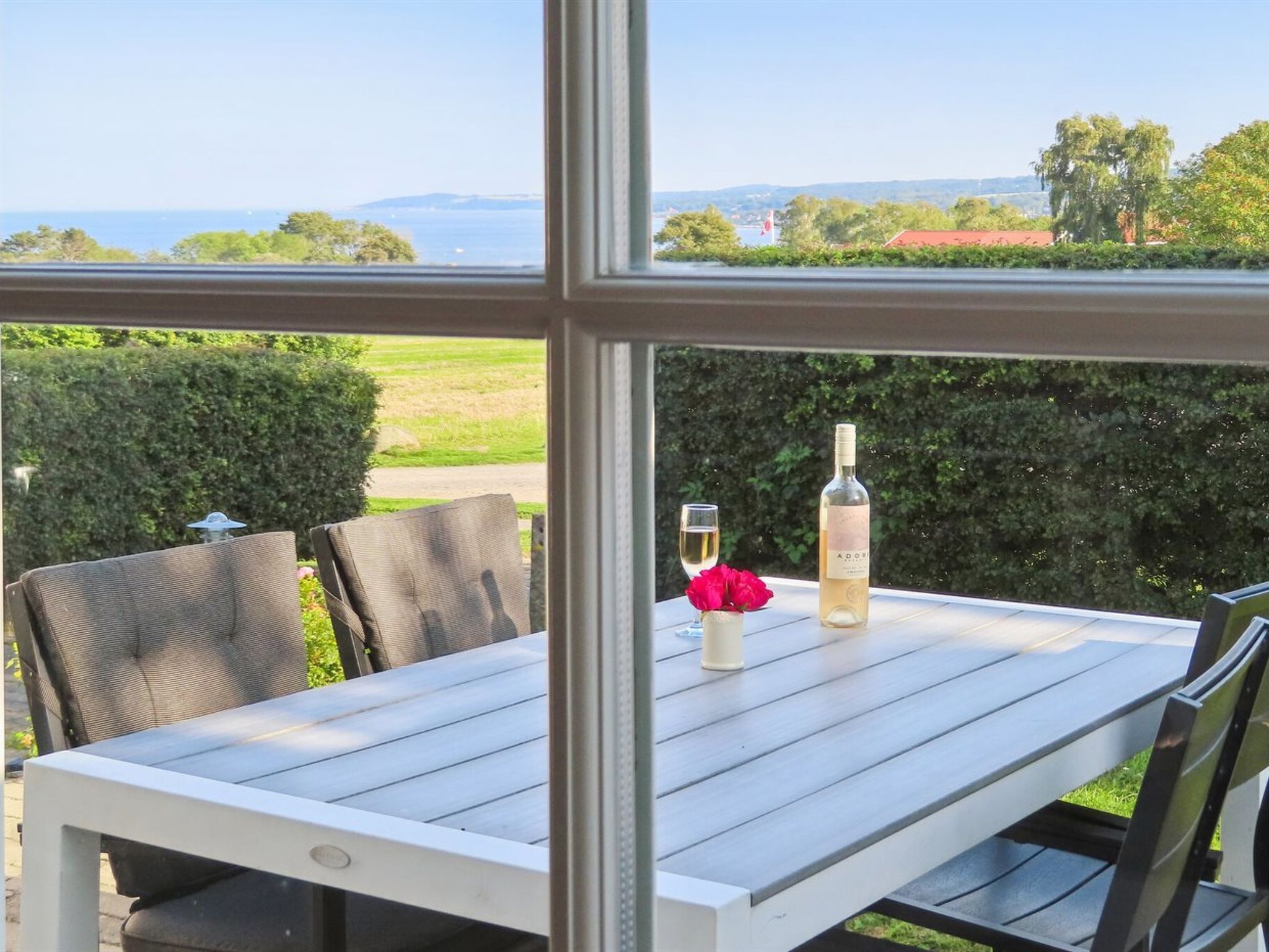 A table with a bottle of wine, a glass, and a rose, set against a view of a green landscape and the sea.