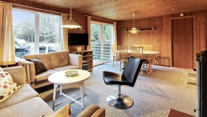 A cozy cabin interior with a wooden ceiling, a sofa, a dining table with chairs, and a TV mounted on the wall.