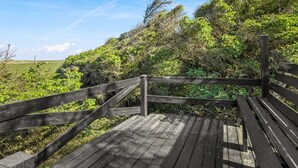 A wooden deck with railings, surrounded by greenery and a clear sky.